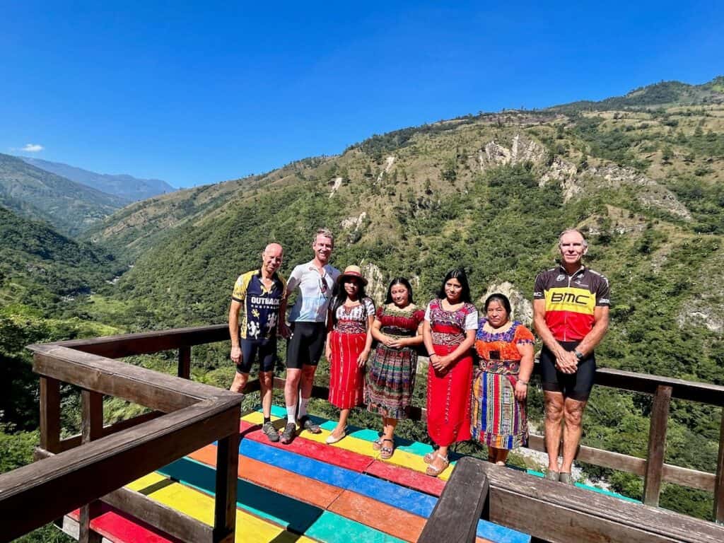 Local women dressed in traditional Guatemalan standing next to foreign cyclists at a beautiful overlook