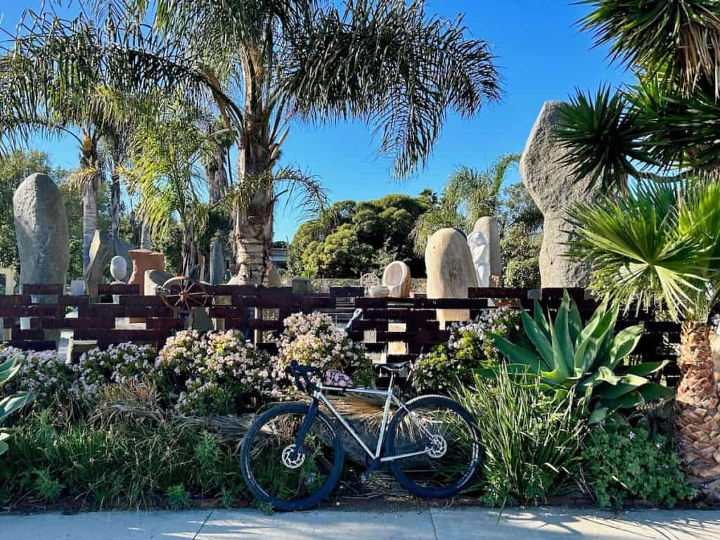 Bike propped up against wall filled with desert plants. Rock sculptures and palm trees in the background