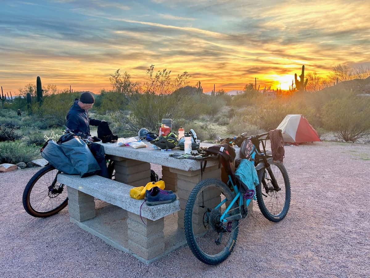Bikepackers unpacking gear at concrete picnic table at campground in the desert at sunset